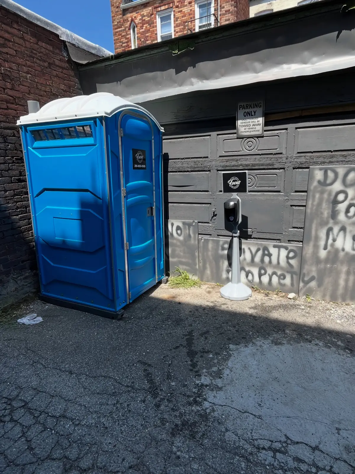 Blue standard porta potty on asphalt beside a garage with a hand sanitizer stand